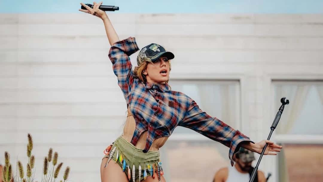 Foto de prensa en vivo de Slayyyter: con gorra, camisa escocesa cortada y un short verde con señuelos de pesca colgando, levanta el micrófono con el brazo estirado frente a una pared de madera blanca. Foto: Joshua Druding