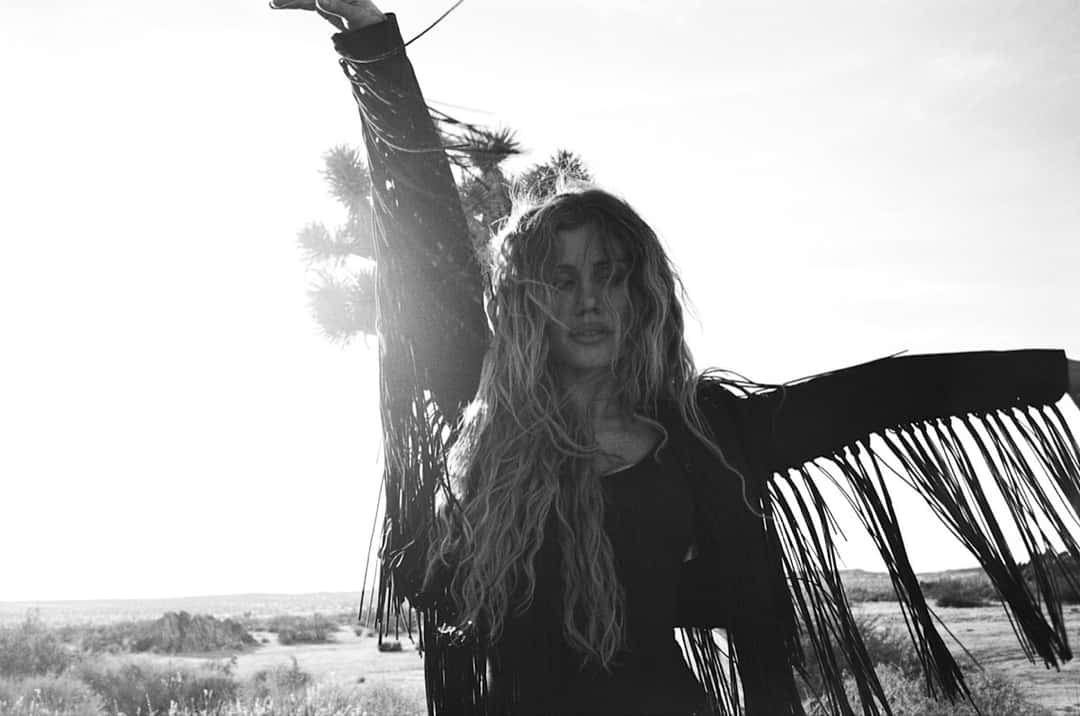 Foto de prensa oficial de Slayyyter en blanco y negro: con una campera de flecos y el pelo largo al viento, levanta un brazo en medio del desierto, con un árbol de Josué detrás y el sol a contraluz