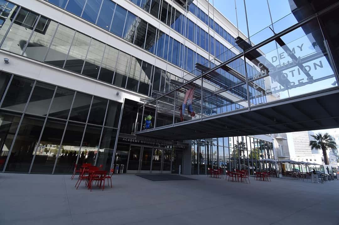 Entrada del Academy Museum of Motion Pictures en Los Ángeles, sobre el Wilshire Boulevard: el edificio Saban con su fachada vidriada, el puente de vidrio con el nombre del museo y mesas rojas en la explanada. Foto: Fred Cherrygarden, Wikimedia Commons, CC BY-SA 4.0