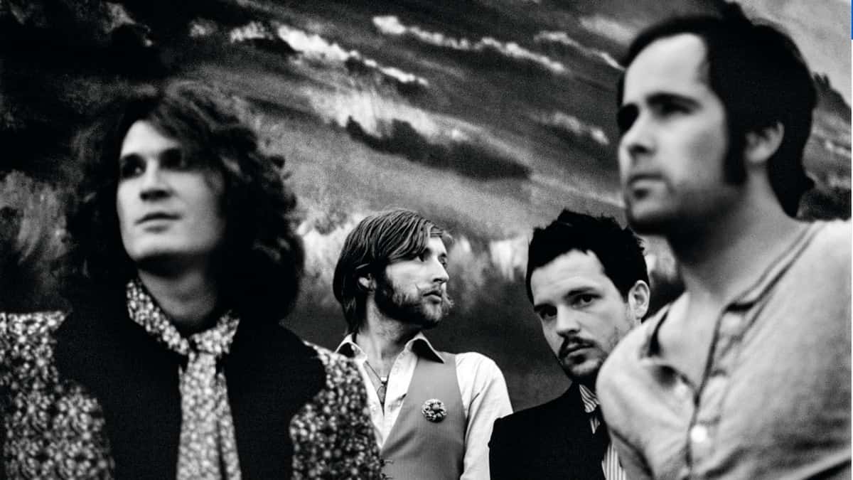 Foto de archivo en blanco y negro de The Killers en la época de Sam's Town: los cuatro integrantes delante de un fondo pintado con un cielo nublado, Brandon Flowers de saco oscuro mirando a cámara