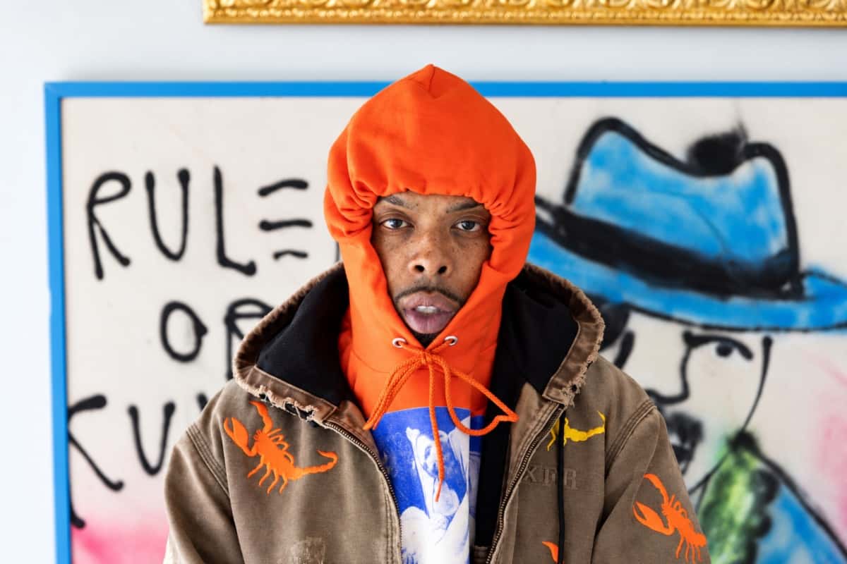 Foto de prensa de Westside Gunn con capucha naranja y campera marrón con escorpiones bordados, frente a una pintura con grafitis