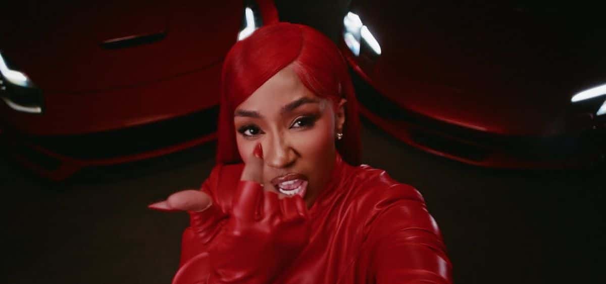 Fotograma del video oficial de «F You»: Yung Miami, con pelo largo rojo, guantes y campera de cuero rojo, mira a cámara y estira la mano hacia adelante, con un auto rojo de fondo