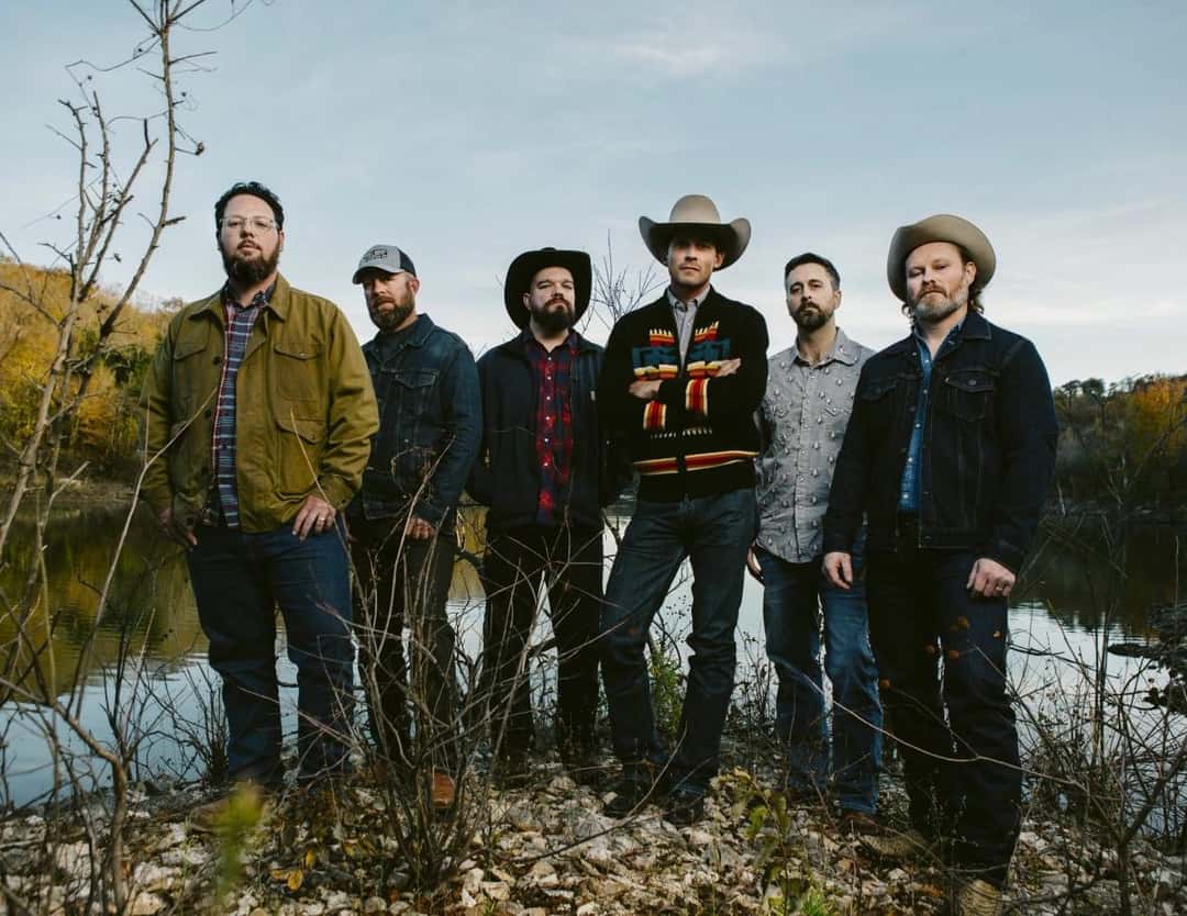 Foto oficial de archivo de Turnpike Troubadours: los seis integrantes de la banda posan de pie a orillas de un lago, con camperas de jean y de trabajo, tres de ellos con sombrero, entre ramas secas y árboles otoñales. Foto: Turnpike Troubadours