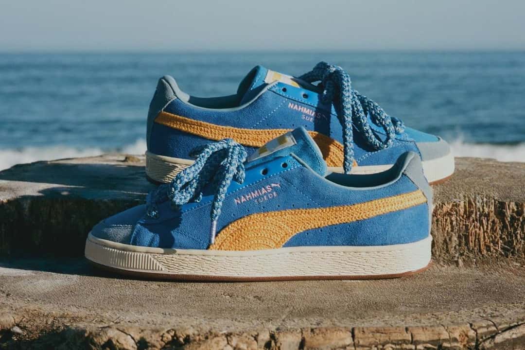 NAHMIAS x PUMA Suede Crochet azul eléctrico sobre un tronco frente al mar, con la Formstrip amarilla tejida y la etiqueta de NAHMIAS en el costado