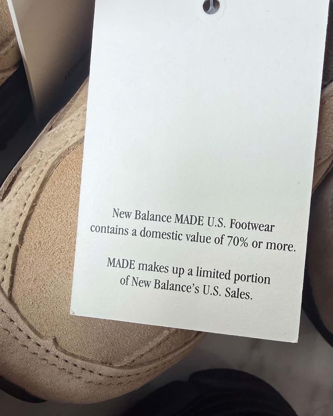 Etiqueta colgante blanca de New Balance MADE U.S. sobre un JJJJound x New Balance 990v3 de gamuza tan, que indica un valor doméstico del 70% o más