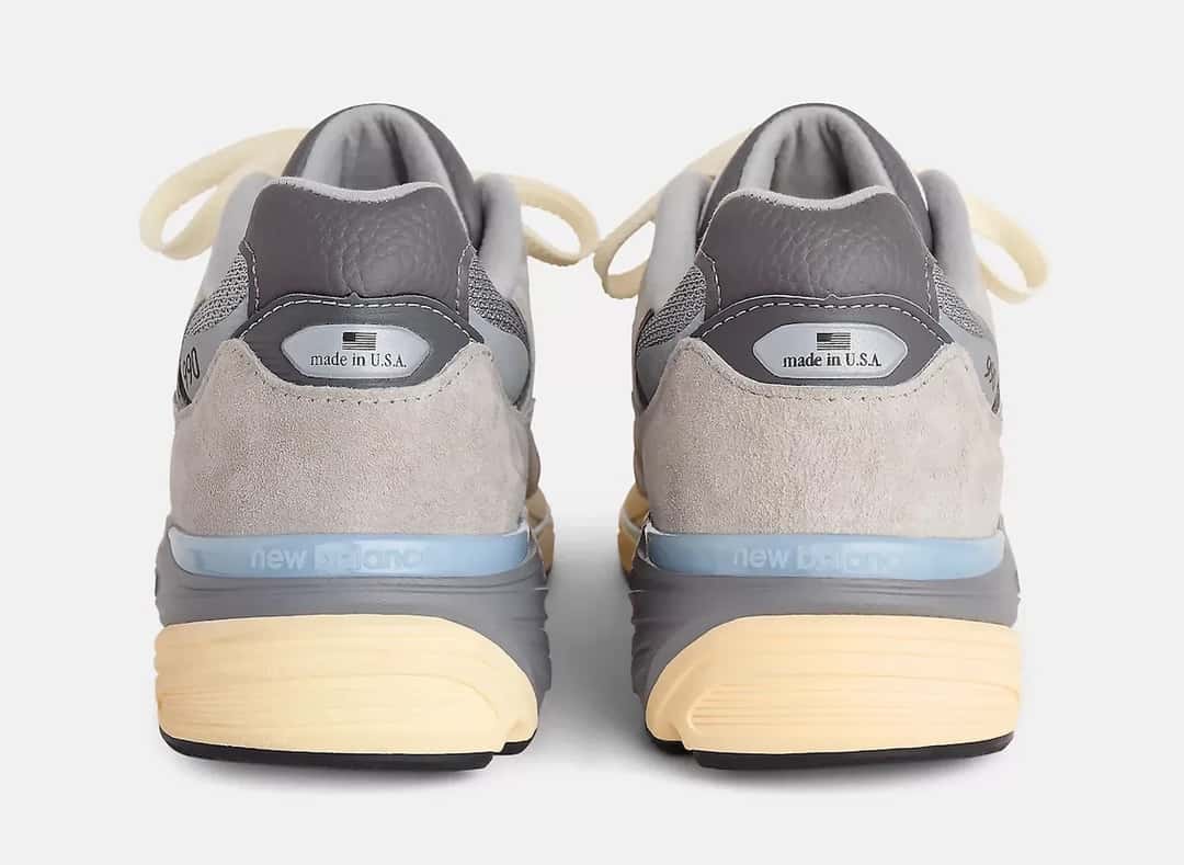 Talones del New Balance 990v7 Made in USA Kool Grey sobre fondo gris claro, con la gamuza gris, la etiqueta Made in USA y la entresuela con una franja celeste y la base amarilla