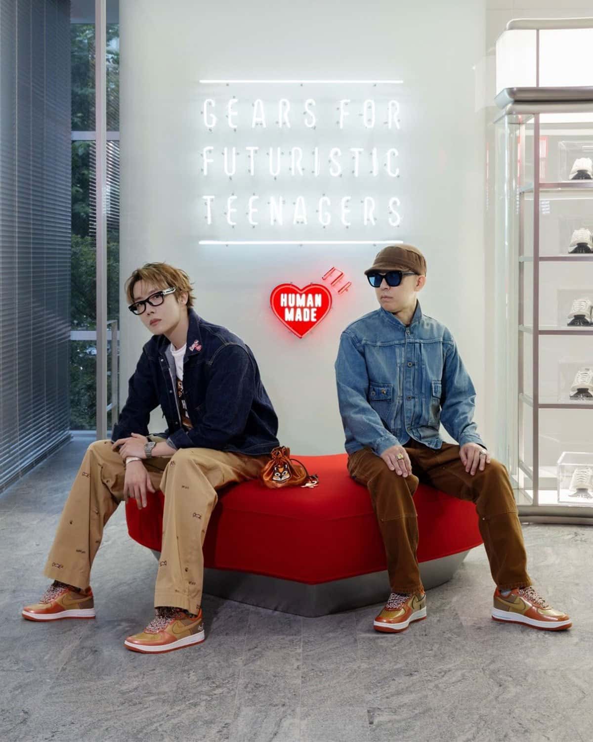 j-hope y NIGO sentados en un banco rojo frente a un cartel de neón de Human Made, los dos con el Nike Air Force 1 '01 Burnt Sunrise en charol cobre y dorado