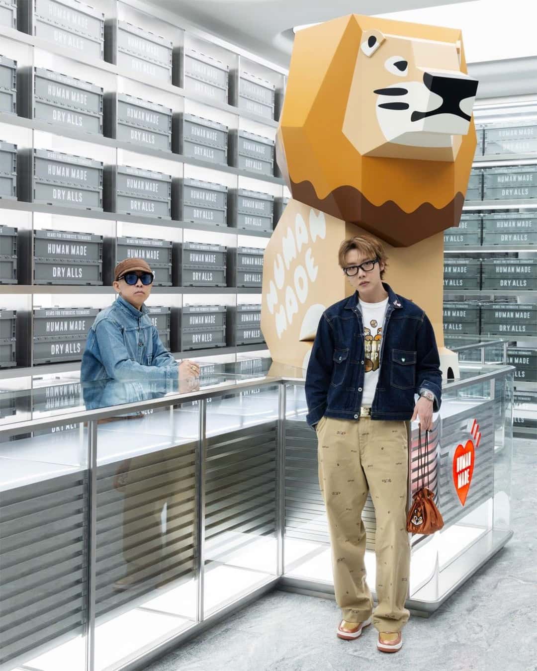 NIGO apoyado en un mostrador de vidrio y j-hope de pie delante de una gran escultura de león de Human Made, con el Air Force 1 Burnt Sunrise en los pies