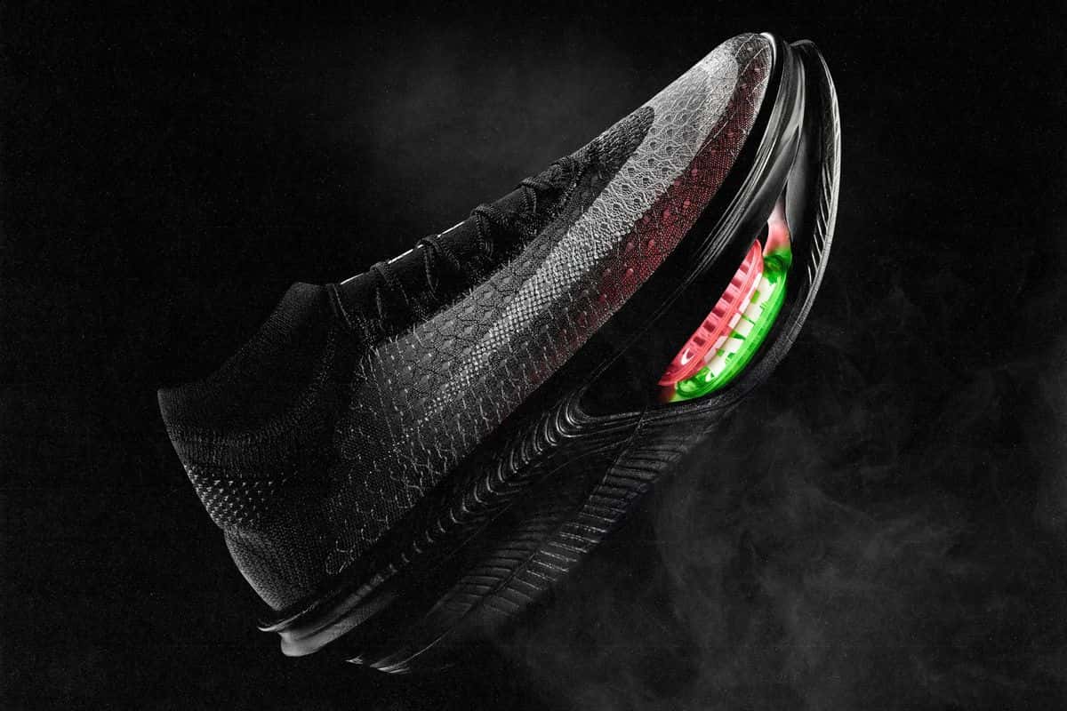 Nike Apex negra de perfil sobre fondo negro con humo, con el upper tejido en tonos grises y rojizos y el doble Air Zoom rojo y verde asomando bajo el antepié