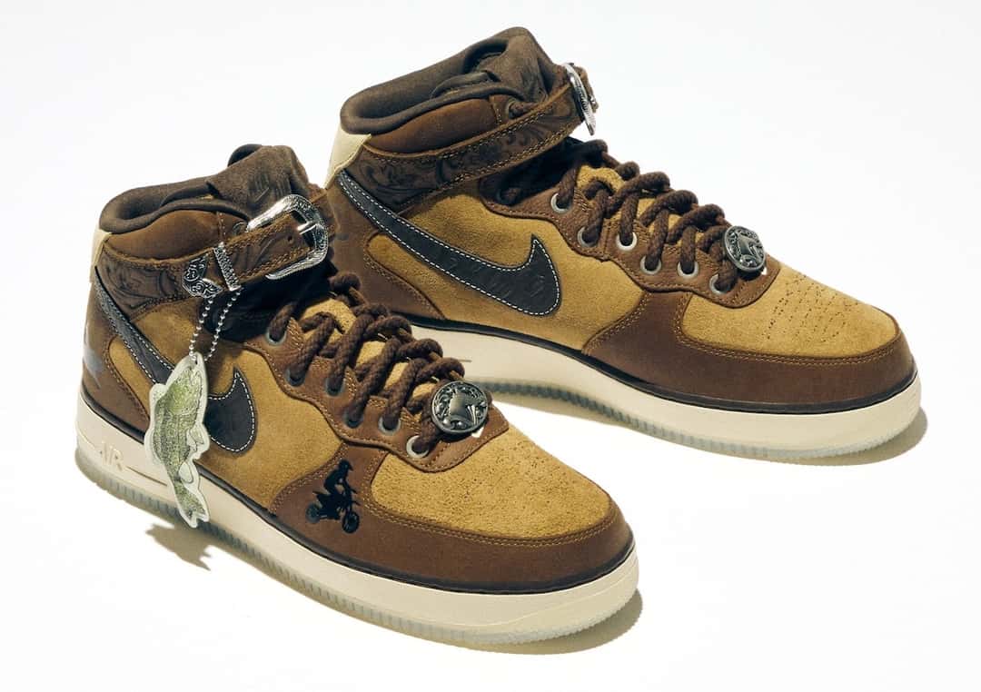 Nike Air Force 1 Mid de la colección Doernbecher Freestyle 22 sobre fondo blanco, en gamuza marrón clara y oscura con swoosh marrón, correa de cuero con hebilla plateada, dijes metálicos en los cordones, un dibujo de un ciclista en la puntera y una etiqueta colgante