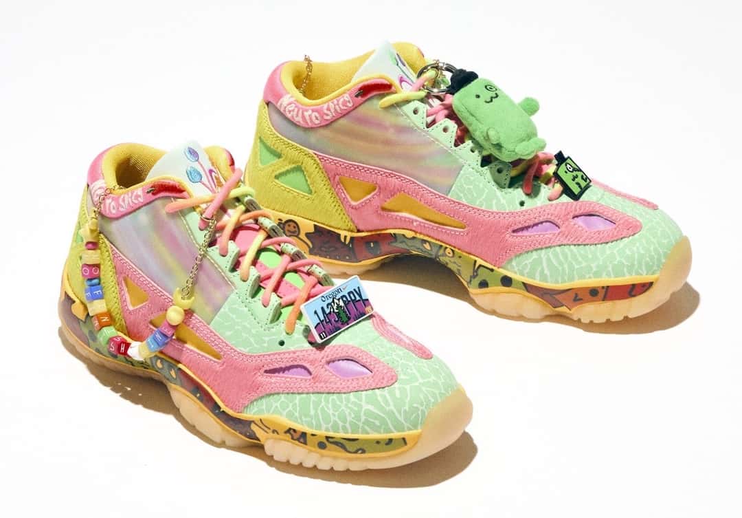 Air Jordan 11 Low IE de la colección Doernbecher Freestyle 22 sobre fondo blanco, con upper verde agua con textura, paneles rosas, zonas tornasoladas, suela translúcida, cordones con cuentas de colores y un muñeco de peluche verde colgado del cuello