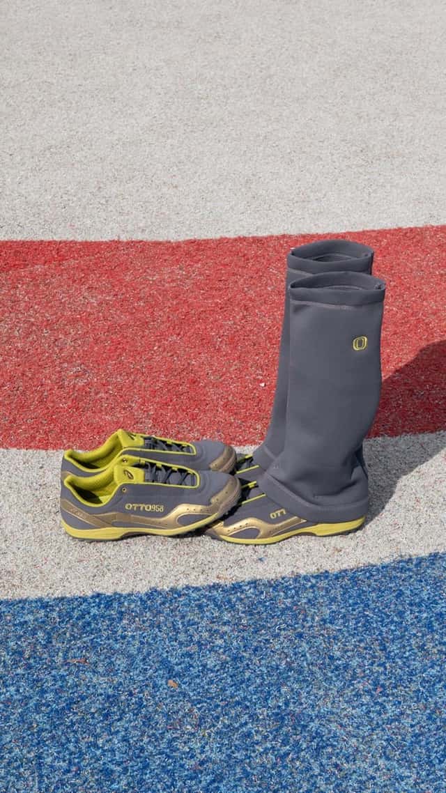 Par de ASICS GEL-BELARRAOTTO de OTTO 958 en gris con detalles verde lima, sobre una pista de colores, con las polainas grises paradas como botas al lado