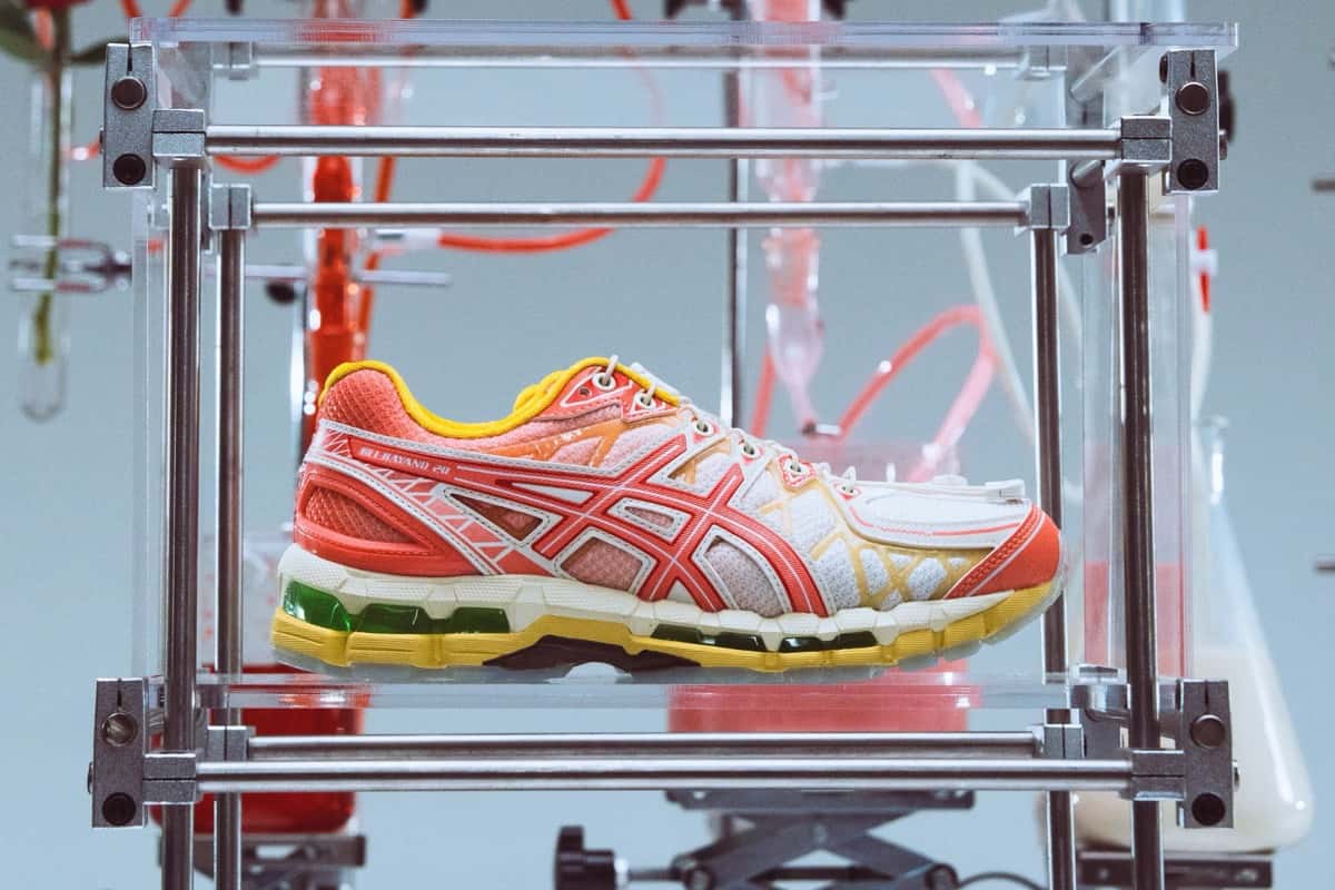 SneakerLAH x ASICS GEL-KAYANO 20 Ice Kacang dentro de una estructura de metal y acrílico, en una escenografía de laboratorio con mangueras rojas y frascos, con upper blanco y coral, cuello amarillo y GEL verde en la mediasuela