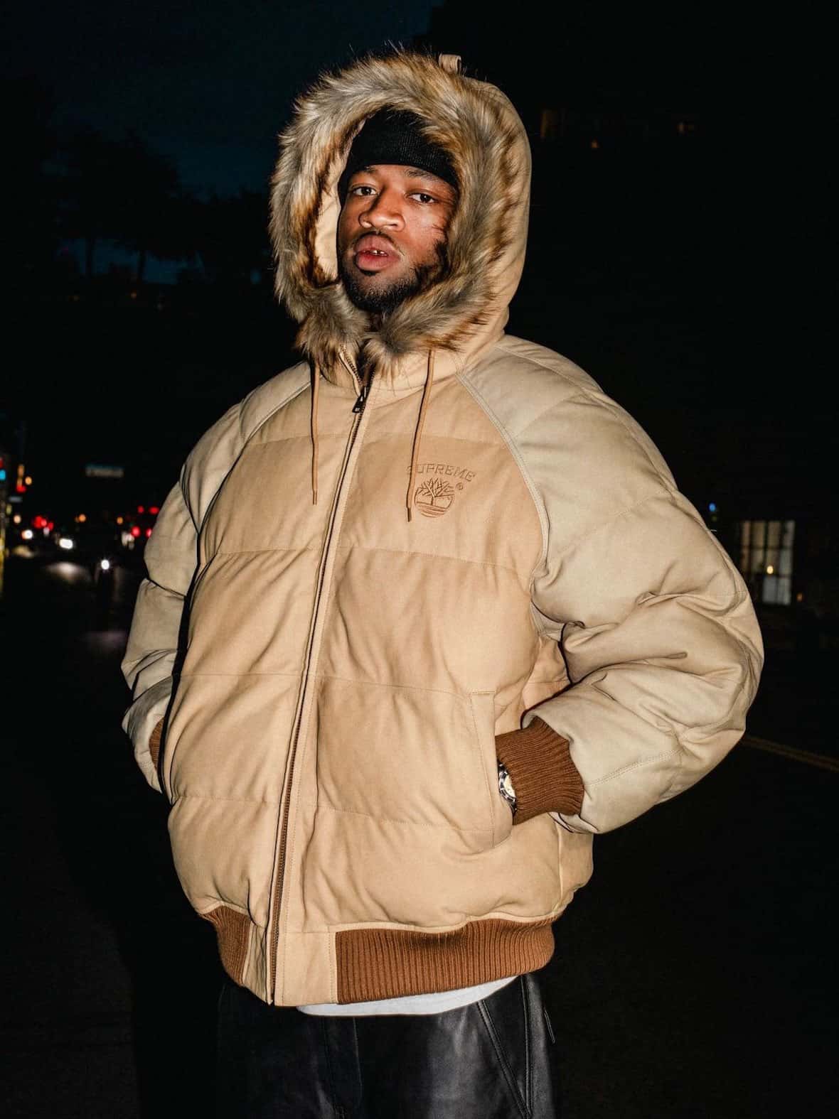 Modelo de noche en la calle con la campera Supreme x Timberland Suede Puffer color wheat, con capucha de piel sintética y gorro oscuro