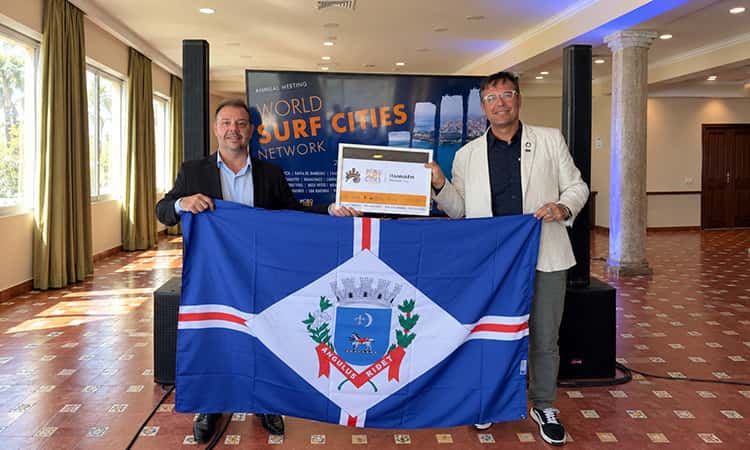 Dos representantes de Itanhaém sostienen la bandera de la ciudad y el certificado de la World Surf Cities Network frente a la pantalla de la asamblea anual en Ericeira