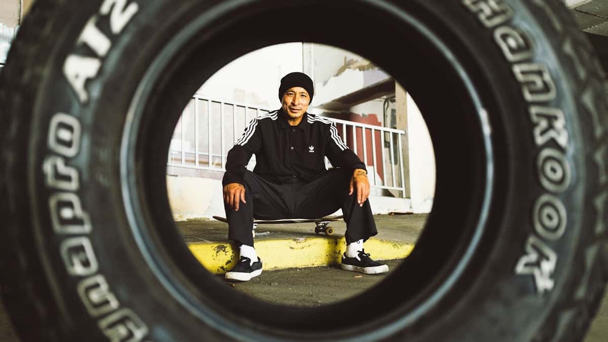 Daewon Song, con gorro negro y campera deportiva negra, sentado sobre su skate en un cordón amarillo, visto a través del hueco de una cubierta de auto