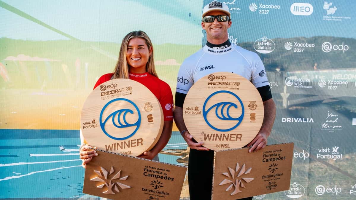 Brisa Hennessy, con la lycra roja, y Crosby Colapinto, con la blanca, sonríen con los trofeos de madera de campeones del EDP Ericeira Pro frente al panel del evento