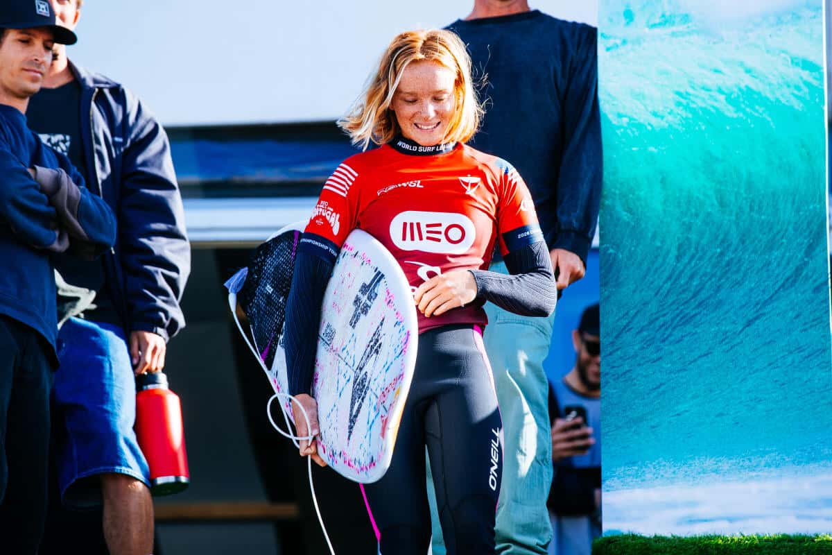 Caitlin Simmers, con la lycra roja del MEO Rip Curl Pro Portugal y su tabla bajo el brazo, sonríe y mira hacia abajo junto a un panel con la imagen de un tubo, en Supertubos