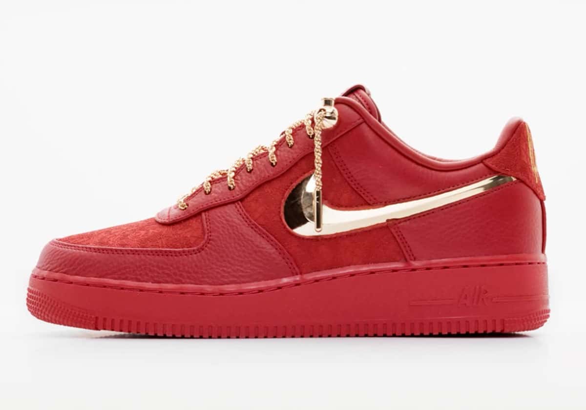 Victor Solomon x Nike Air Force 1 Low Trophies rojo de perfil sobre fondo blanco, con el Swoosh dorado metálico en relieve, cordones de soga dorados con un dije colgante y suela roja