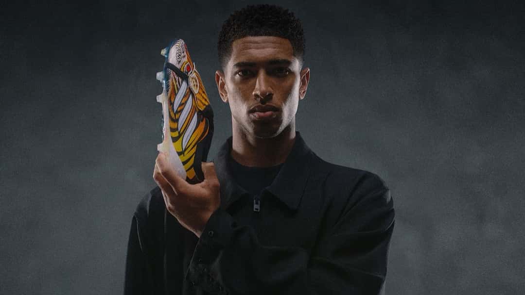 Un futbolista con campera negra sostiene la adidas Predator Y-3 con el tigre pintado, sobre fondo gris oscuro